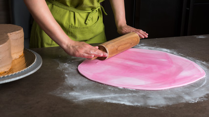 Woman using rolling pin preparing pink fondant for cake decorating, hands detail
