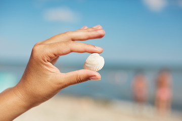 Little beautiful sea shell in woman`s hands against beutiful marine background. Woman having holiday on beach, demonstrating wonderful sea shell which she found on sand beach. Summer and vacation