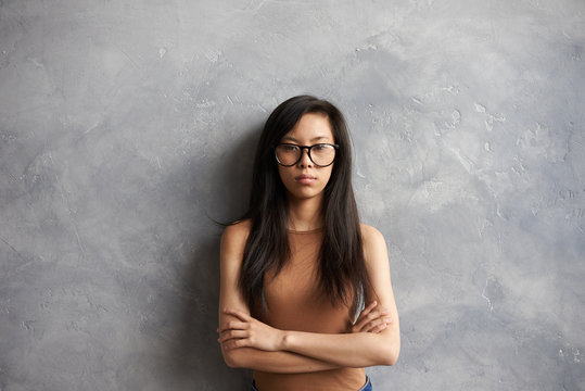 Isolated Waist Up Portrait Of Serious Young Brunette Woman Of Asian Appearance Wearing Glasses Posing At Blank Wall With Arms Crossed, Feeling Unhappy Or Angry After Fight With Her Boyfriend