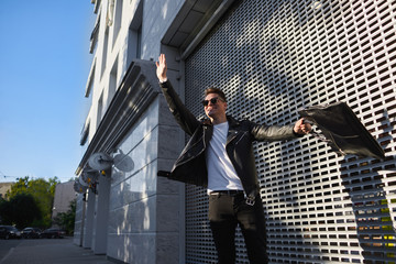 Outdoor lifestyle picture of handsome guy in shades standing on street holding bag in one hand and raising the other, waiving it actively, hailing a cab while being late to airport or urgent meeting