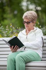 Senior grandmother reading black book while sitting on the bench in park