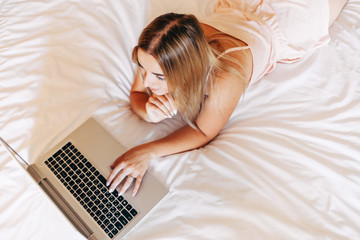 Young smiling woman in pajamas on white bed working on a laptop