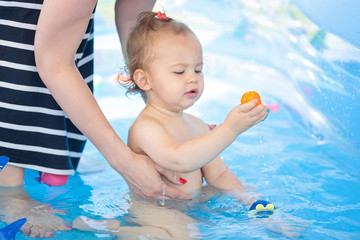 Sixteen months old baby girl playing in the plastic pool with toys - mother standing next to her, keeping an eye and watching - baby covered with sun protection cream; happy childhood concept