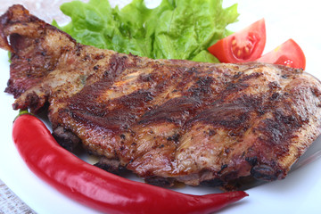 Pork ribs on grill and chili pepper with tomato, lettuce leaves on white plate