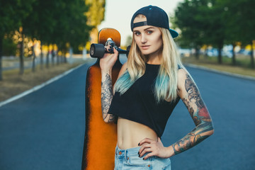 A beautiful, sexy hipster blonde with blue hair in tattoo stands with a longboard  on the street.