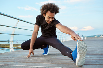 Dark-skinned man athlete in black sportswear and blue sneakers stretching his legs with lunge hamstring stretch exercise on the pier. Afro-American young male runner warming-up his muscles before