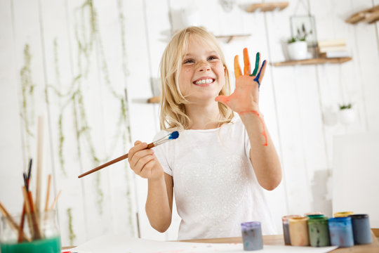 Happy and playful cute freckled blonde girl dressed in white, holding brush in one hand and showing another hand, which she messed up with paint. Children and happiness concept. Isolated shot