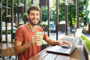 View of glad caucasian bearded man using laptop computer smiling with teeth, surfing internet sitting at wooden table at summer cafeteria and drinking lemonade.