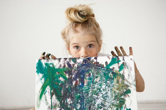 Close-up portrait of blonde European little girl with hair bun and big blue eyes demonstrating her picture. Talented kid makes parent feel good. Child is a source of positive emotions.
