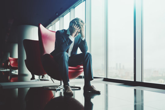 Sad businessman is sitting on red armchair and mourning for his dismissal after conversation with boss; man entrepreneur has just known about bankruptcy of his company and grieving over it