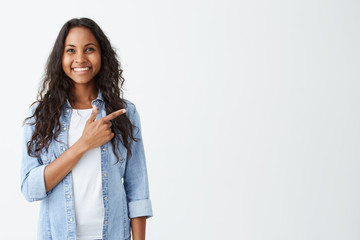 Fashionable emotional young African American female wearing denim shirt pointing her index finger at white blank wall background behind her, looking positive and happy, broadly smiling.