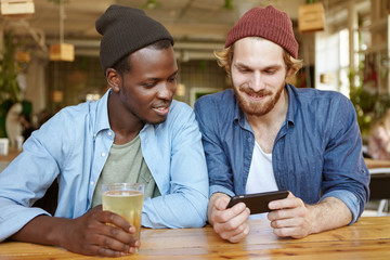 Young hipster guy with ginger beard, holding cell phone in hands showing his favourite trailer to his dark-skinned friend while sitting at pub and drinking cold drinks. Interracial men`s friendship