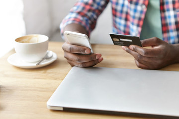 Close up shot of black man's hands holding credit card and mobile phone over wooden cafe table with coffee cup and laptop computer. Technology, internet banking, e-commerce and online trading concept