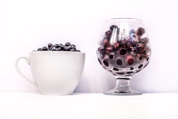 white Cup with black blueberries and transparent glass with frozen currants on white background