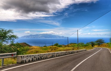Cloudscape over asphalt road heading down towards the capital city of São Filipe, in Fogo, Cabo Verde, with the island of Brava sitting at sea.