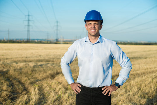 engineer or worker smiles in protective helmet holding a phone. Wheat fiels, power line, sunny day, blue sky.