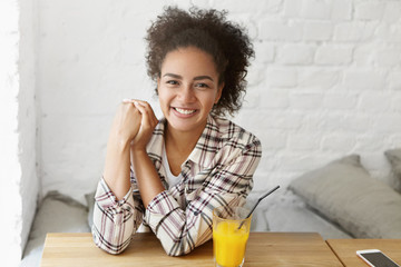 Portrait of positive smiling young mixed-race woman with Afro hairstyle relaxing at cozy cafe, having fresh orange drink, sitting on comfortable couch by the window, her cell phone resting on table