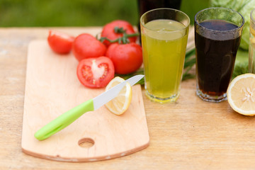 Yellow fresh lemon, red tomato and steel knife on the wooden cutting Board on a background of fresh vegetables and glass cups with a summer drink.
