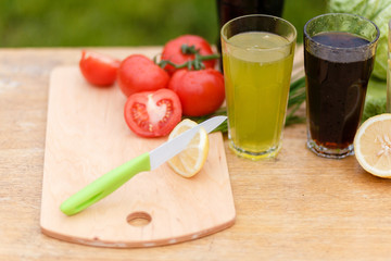 Yellow fresh lemon, red tomato and steel knife on the wooden cutting Board on a background of fresh vegetables and glass cups with a summer drink.