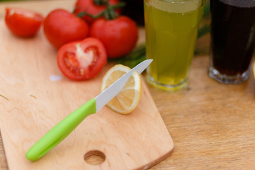 Yellow fresh lemon, red tomato and steel knife on the wooden cutting Board on a background of fresh vegetables and glass cups with a summer drink.