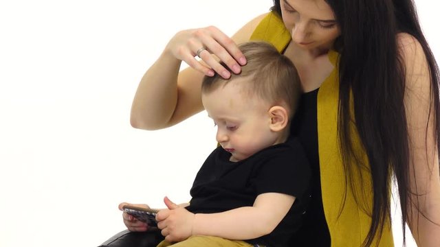 Woman is sitting on the floor with her baby, she is stroking his hair. White background