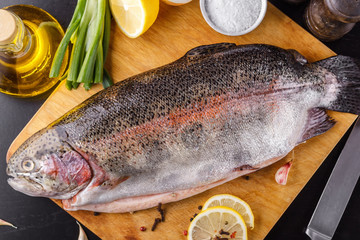 Fresh gutted trout on a wooden cutting board surrounded by ingredients for baking: olive oil, lemon, spring onions, garlic and spices. Top view