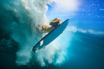 Slim blonde girl in bikini a surfer with surf board duck diving underwater under ocean reflected...