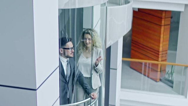 Tilt up of cheerful businessman in glasses wearing suit and businesswoman with blond curly hair chatting in moving glass elevator in modern multi-storey business centre 