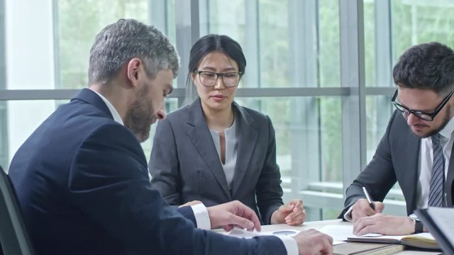 PAN of female Asian corporate lawyer giving contracts to bearded businessmen in suits for signing, then smiling and observing them shaking hands in agreement