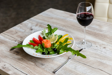 A white plate with salad, that contains salad leaves, fresh salted red fish, tomatoes, black olives, yellow pepper and lemon. Knife and fork. Glass of red wine. Selective focus.