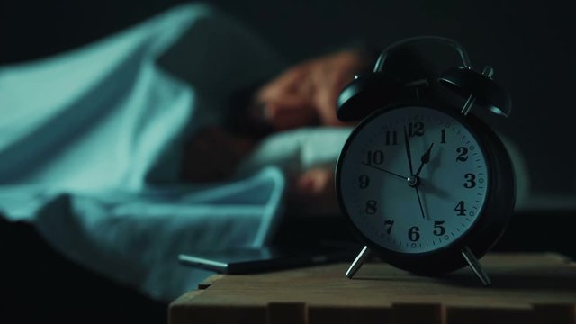 Adult caucasian man talking on mobile phone late at night while lying in bed in bedroom, selective focus on alarm clock on bedside table