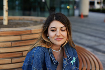 Fototapeta premium Close up shot of gorgeous young Caucasian female wearing jeans jacket, earrings and nose-ring relaxing on wooden bench outdoors, looking down with cute shy smile. Urban style and fashion concept