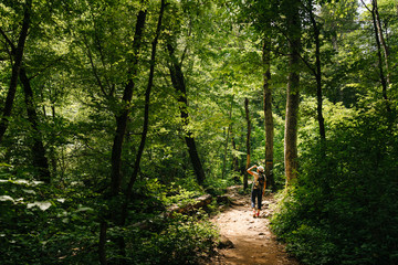 middle-aged woman with a backpack and wearing a hat walks along a forest path. The man on the walk is illuminated by the sun.