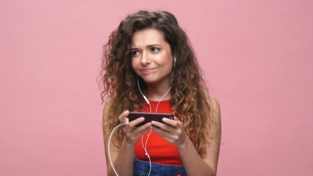 Young amazing caucasian woman talking by earphones and watch video isolated over pink background