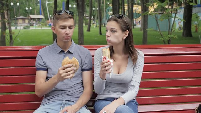 Couple sitting on a red bench in a city park with relish eating a hamburger and hot dog