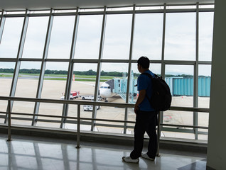Man looking on the private jet,Asia young man is standing near window at the airport and watching plane before departure. He is standing and carrying luggage. Focus on his back,tourist,trip.