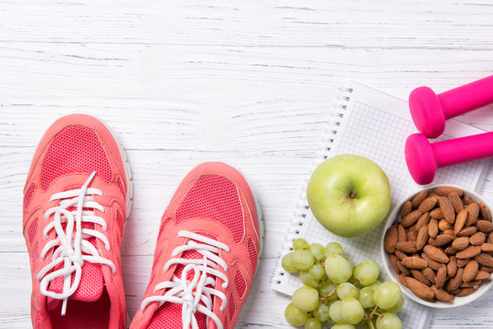 Fitness and healthy eating concept, pink sneakers and dumbbells with apple, grapes and almond nuts on notepad, wooden background, top view with copy space