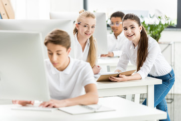 smiling teenage girls looking at camera while sitting at table in classroom with classmates