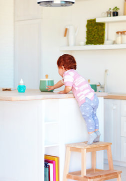 cute toddler baby climbs on step stool, trying to reach things on the high desk in the kitchen