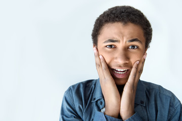 Fototapeta na wymiar portrait of shocked african american teen boy with hands on cheeks looking at camera isolated on white