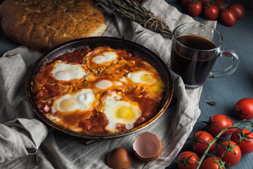 Shakshuka with pita bread in a pan. Fried eggs, onion, bell pepper, tomatoes and parsley on a rustic wooden table with ingredients.