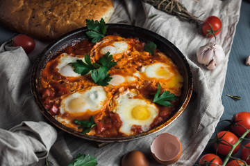 Shakshuka with pita bread in a pan. Fried eggs, onion, bell pepper, tomatoes and parsley on a rustic wooden table with ingredients.