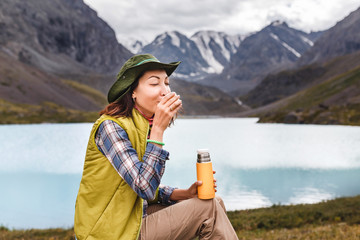 Hiker woman drinks hot tea from a thermos on the shore of a mountain blue lake