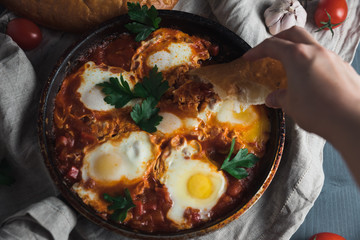 Shakshuka with pita bread in a pan. Fried eggs, onion, bell pepper, tomatoes and parsley on a rustic wooden table with ingredients.
