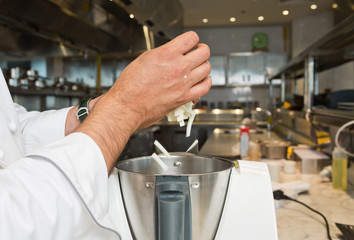 A hand dropping freshly cut slices of celeriac into a food processor, in an industrial kitchen setting.