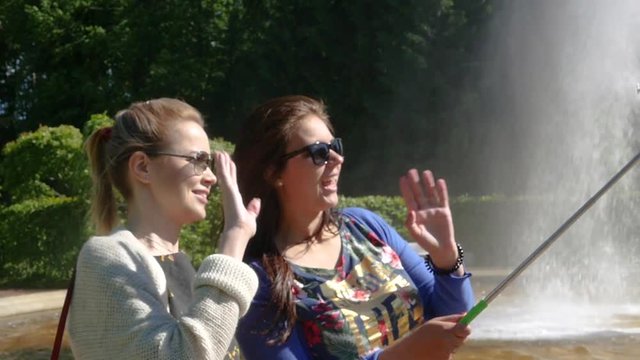 Girlfriends talking on a video call waving their hands at the screen in a green park near the fountain. slow motion. 1920x1080. full hd