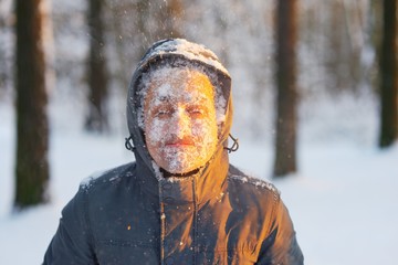 Fototapeta na wymiar Frozen young man in a jacket with a hood, covered with snow in winter forest during sunset. Snowflakes swarmed all over the face