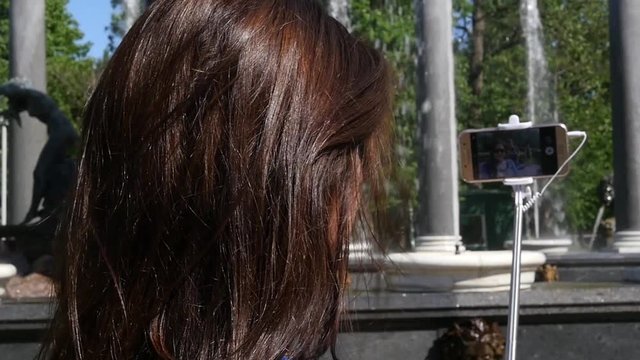 Young girl makes a photo on the phone and a selfie stick, on a background of a fountain in the park, view from behind. slow motion. 1920x1080. full hd