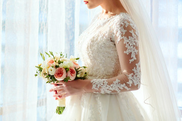 A beautiful bride is holding a wedding bouquet with white roses and peach peonies on a bright window background. Close-up