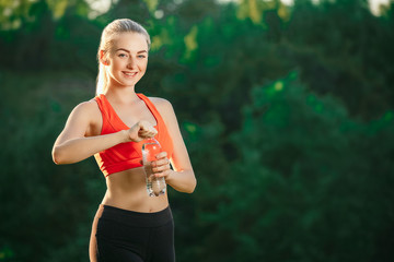 A young blonde in a red top and black pants holds a bottle of water in her hands and opens it to drink in nature.